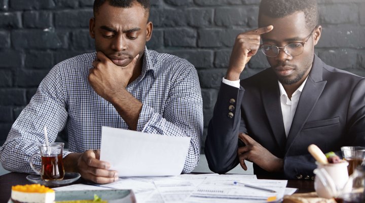 Two worried serious Afro-American businessmen working through papers and discussing financial health report having concentrated looks, sitting close to each other against black brick wall background